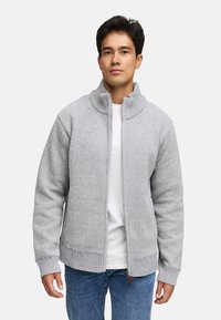 Grå strikket sweater med høj krave og fuld lynlås, med ribbede kanter og en struktureret stof. Bæres over en hvid skjorte og blå jeans.