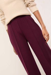 Pantaloni burgundii cu o croială ajustată, având o textură netedă, pliuri frontale și un talie înaltă. Pulover cremos confortabil răsărind deasupra.