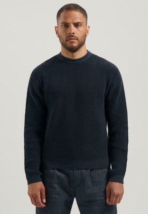 Marineblauer Strickpullover mit strukturiertem Muster, runder Halsauschnitt, gerippte Bündchen und Saum. Kombiniert mit dunkelgrauen Hosen.