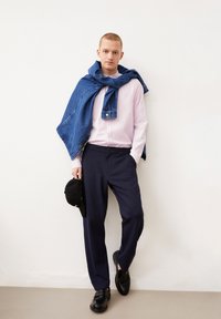 Lichtroze overhemddas, donkerblauwe broek, spijkerjack over de schouders gedrapeerd, zwarte pet in de hand, en zwarte schoenen.