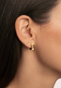 Pendientes de oro con un diseño geométrico. Un pendiente es un grueso aro con un acabado brillante, el otro es un pequeño cuadrado con una piedra clara.