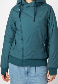 Veste à capuche zippée couleur sarcelle avec cordons et poignets côtelés portée sur un jean bleu clair, montrée du cou aux hanches.