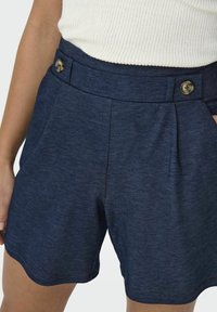 Shorts bleu marine en tissu texturé, avec une taille haute, des plis sur le devant et deux boutons décoratifs à la taille.
