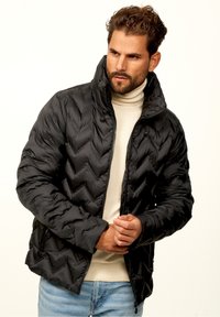 Black puffer jacket with a chevron quilted pattern, high collar, zip closure, and side pockets, paired with a cream turtleneck underneath.