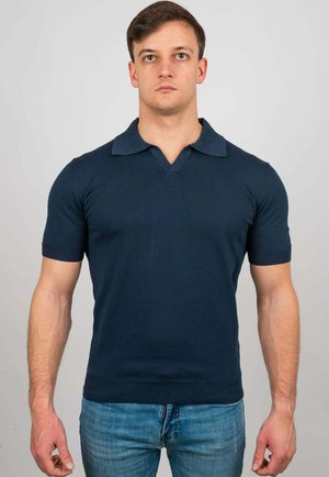 Uomo che indossa una polo blu navy aderente a maniche corte e jeans blu, in piedi davanti a uno sfondo chiaro e uniforme.