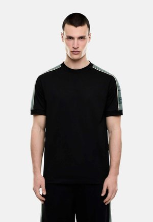 Giovane uomo con capelli corti che indossa una maglietta nera con strisce chiare sulle spalle e una toppa sulla manica sinistra, in piedi contro uno sfondo bianco.