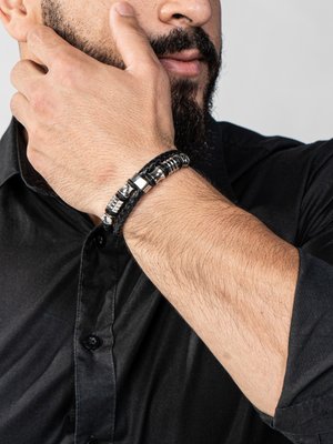 Schwarzes, geflochtenes Lederarmband mit silbernen Akzenten und zylindrischer metallischer Hardware, getragen an einer Hand vor einem dunklen Hemd-Hintergrund.