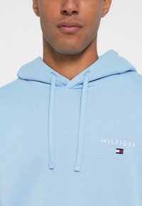 Ανοιχτό μπλε φούτερ από μαλακό ύφασμα. Διαθέτει μπροστινή τσέπη, ρυθμιζόμενη κουκούλα με κορδόνι και κεντητό λογότυπο σε λευκές και κόκκινες λεπτομέρειες.