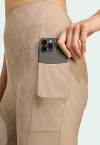 Beige Leggings mit dezentem Muster, ausgestattet mit einer Seitentasche, in der ein Smartphone steckt. Reflektierendes Logodesign an der Hüfte und glatte Textur.