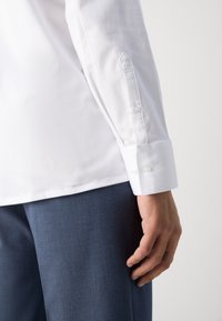 Biała bawełniana koszula z zapięciem na guzik w mankietach, z wyraźnym, spiczastym kształtem mankietów. Granatowe spodnie częściowo widoczne. Gładka tekstura.