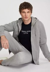 Szara bluza z kapturem zapinana na zamek i dopasowane spodnie dresowe, pod spodem czarna koszulka z grafiką. Miękka tkanina i luźny krój. Prosty design.