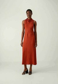 Nő, aki ujjatlan, rozsdabarna midi ruhát visel, cseppnyakú, fekete szíjú, tömbsarkú szandálokkal, egy egyszínű világos háttér előtt áll.