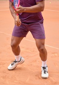 Ljubičaste sportske kratke hla�če s Fila logotipom, uparene s bijelim čarapama do gležnja i pretežno bijelim teniskim cipelama, na narančastom zemljanom terenu.