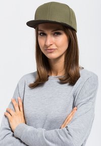 Cappellino da baseball verde oliva con visiera piatta, realizzato in tessuto testurizzato. Indossato con un maglione grigio chiaro, che mostra un fit rilassato e informale.