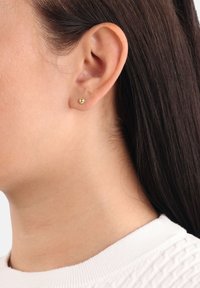 Guldstiftörhänge som visas på en modells öra, med en slät, sfärisk design och en glänsande yta. Håret är mörkt och rakt.