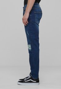 Jean en denim bleu foncé avec une coupe slim, présentant une zone distressed au genou et une taille standard. Associé à des chaussures Vans noires.