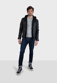 Mladý muž stojící, oblečený v černé kožené bundě, šedém svetru, tmavě modrých džínách s ohrnutými nohavicemi a černo-bílých teniskách na jednobarevném světlém pozadí.