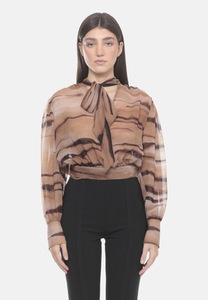 Giovane donna con capelli scuri e lisci che indossa una camicetta marrone trasparente a righe con fiocco e pantaloni neri a vita alta, in piedi davanti a uno sfondo bianco.