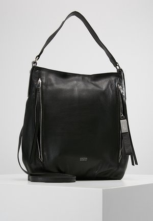 Bolso de cuero negro con correa para el hombro, dos bolsillos frontales verticales con cremallera y una pequeña etiqueta metálica con el logotipo sobre un pedestal blanco.