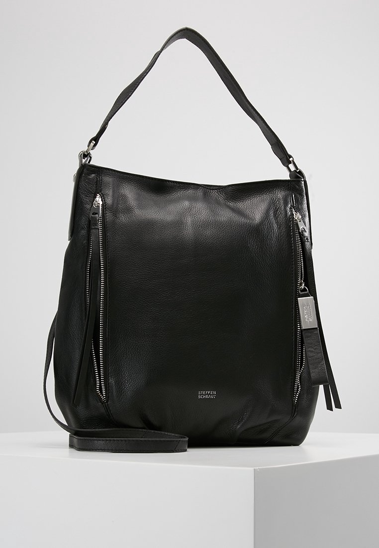 Sac à main en cuir noir avec bandoulière, deux poches zippées verticales à l'avant et une petite plaque avec logo en métal sur un socle blanc.
