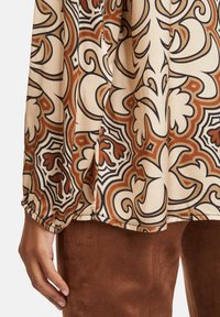 Långärmad blus i ljusbeige med utsmyckade bruna och svarta blommönster, med elastiska ärmslut och en sidospalt.