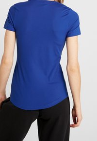Top ajustado de manga corta en un material azul brillante. Textura suave con un cuello redondo y dobladillo curvado, combinado con pantalones negros.