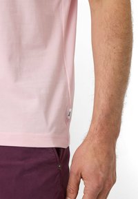 T-shirt in cotone rosa chiaro con scollo rotondo, maniche corte e etichetta laterale. Abbinato a pantaloni viola scuro, mostranti un tessuto текстurizzato.