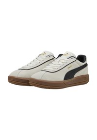 Baskets en suede blanches avec des accents noirs et une semelle en caoutchouc marron. Elles disposent de lacets blancs et d'un logo doré sur le côté et la languette.
