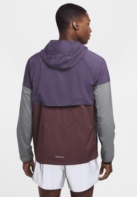 Chaqueta con capucha que presenta un diseño a bloques de color en púrpura, gris y marrón; material ligero, puños elásticos y un detalle de bolsillo trasero.
