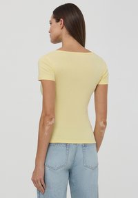 Maglietta gialla a maniche corte, aderente, con una texture morbida e scollo a barca; abbinata a jeans chiari a vita alta, con dettagli di cucitura visibili.