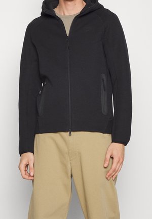 Man draagt een zwart hoodie met ritssluiting, capuchon en ritssluitingen aan de zijkanten, gecombineerd met beige broek en een lichtbruin shirt.