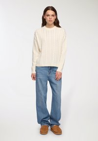 Biały sweter w warkocze z okrągłym dekoltem, noszony z jasnoniebieskimi, luźnymi dżinsami oraz brązowym obuwiem wsuwanym. Miękka faktura, codzienny design.