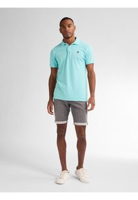 Camisa polo azul claro de mangas cortas, con logotipo en el pecho, combinada con pantalones cortos grises. El modelo lleva zapatillas blancas y está de pie sobre un fondo neutro.