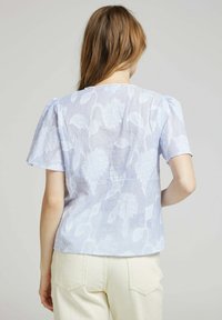 Blouse bleu clair avec un motif floral ton sur ton, manches courtes bouffantes et col rond. Fabriquée en tissu léger et texturé.