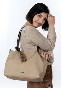 Sac cabas en toile beige avec des accents en cuir marron, doté d'un pompon décoratif et d'une plaque logo sur le devant. Design grand et spacieux.