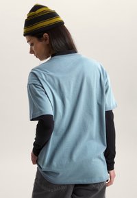 Camiseta de algodón azul claro con mangas cortas, corte relajado y ligera textura, llevada sobre una camiseta de manga larga negra. Gorro a rayas amarillo y negro.