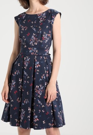 Femme portant une robe sans manches bleu marine avec un motif floral et papillon et une jupe plissée.