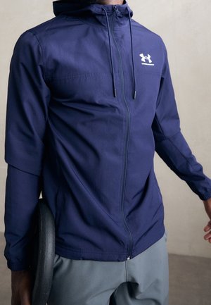 Mężczyzna w granatowej bluzie Under Armour na zamek, trzymający czarną ciężarkę po boku, widoczne szare spodnie.