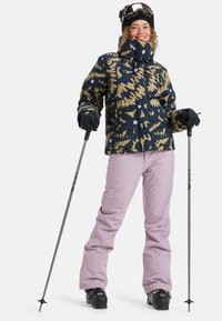 Skijack met een marineblauw en beige abstract patroon, hoge kraag en knopen. Gecombineerd met lichtroze sneeuwbroeken en zwarte skischaatsen.