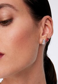 Złote kolczyki z przezroczystymi prostokątnymi i głęboko czerwonymi ovalnymi kamieniami szlachetnymi, o eleganckim, nowoczesnym wzornictwie, noszone na uchu modelki.