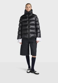 Femme portant une doudoune noire brillante, un short noir arrivant aux genoux, des chaussettes bleu marine montantes et des mocassins noir et blanc, debout devant un fond uni.