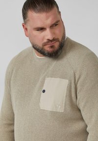 Beżowy sweter z dzianiny o teksturowanym wzorze, z jasnobeżową kieszonką na lewym boku ozdobioną niebieskim guzikiem. Okrągły dekolt.