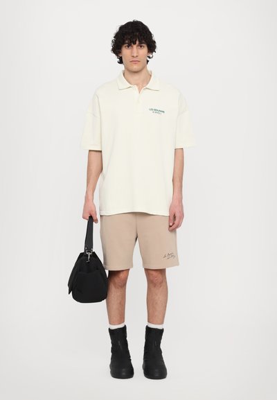 Polo shirt crème avec texte vert, shorts beiges avec couture accentuée, sac à bandoulière noir et bottes zippées noires avec doublure blanche.