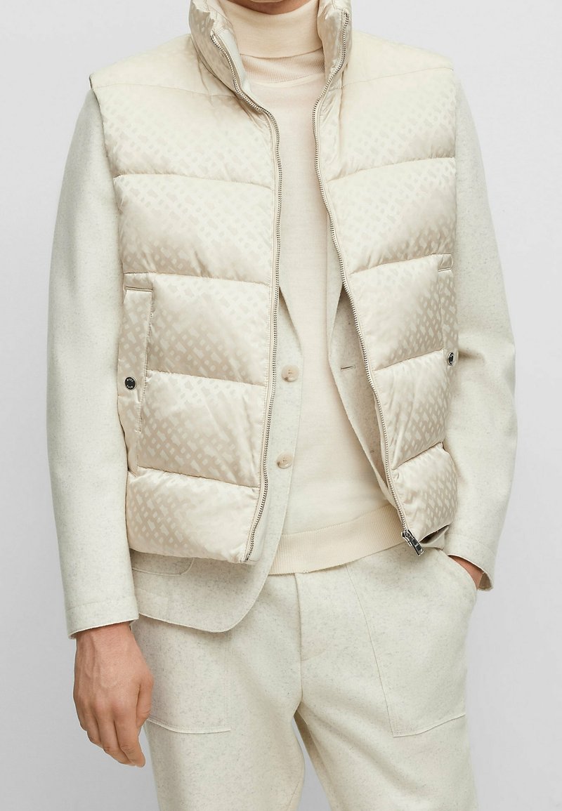 Puffer vest in light beige, featuring a subtle geometric pattern, high collar, and zip closure. Worn over a beige sweater and tailored pants.