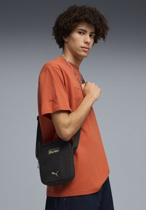 Zwarte crossbodytas met gestructureerde stof, gouden merkopdruk en verstelbare riem, gedragen over een oranje shirt met reliëflogo.