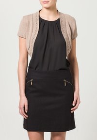 Beige öppen stickad kortärmad cardigan över en svart blus och figurnära svart kjol med dragkedje-fickor, slät textur, strukturerad design.