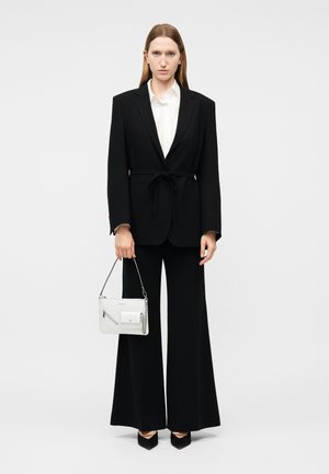 Mujer con pantalón ancho negro y camisa blanca sosteniendo un bolso blanco, de pie sobre un fondo blanco liso.