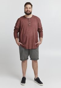 Camicia bordeaux a maniche lunghe con scollatura a bottoni, abbinata a pantaloni corti grigi. Sneakers nere completano l'outfit.