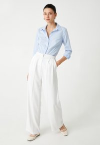 Camisa de botón azul claro de manga larga, combinada con pantalones anchos blancos de talle alto y zapatos planos beige. Diseño simple y limpio.