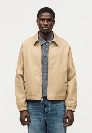 Man wearing beige zip-up jacket over blue patterned polo shirt and blue jeans, standing against a plain light background.